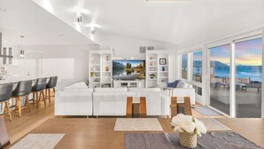 Una sala de estar moderna con un sofá blanco grande, un comedor con mesa de madera y vistas al mar a través de amplios ventanales.