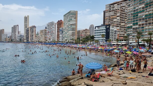 A crowded beach with people swimming and sunbathing, surrounded by tall buildings.