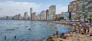 En fullsatt strand med folk som badar och solar, omgiven av höga byggnader.