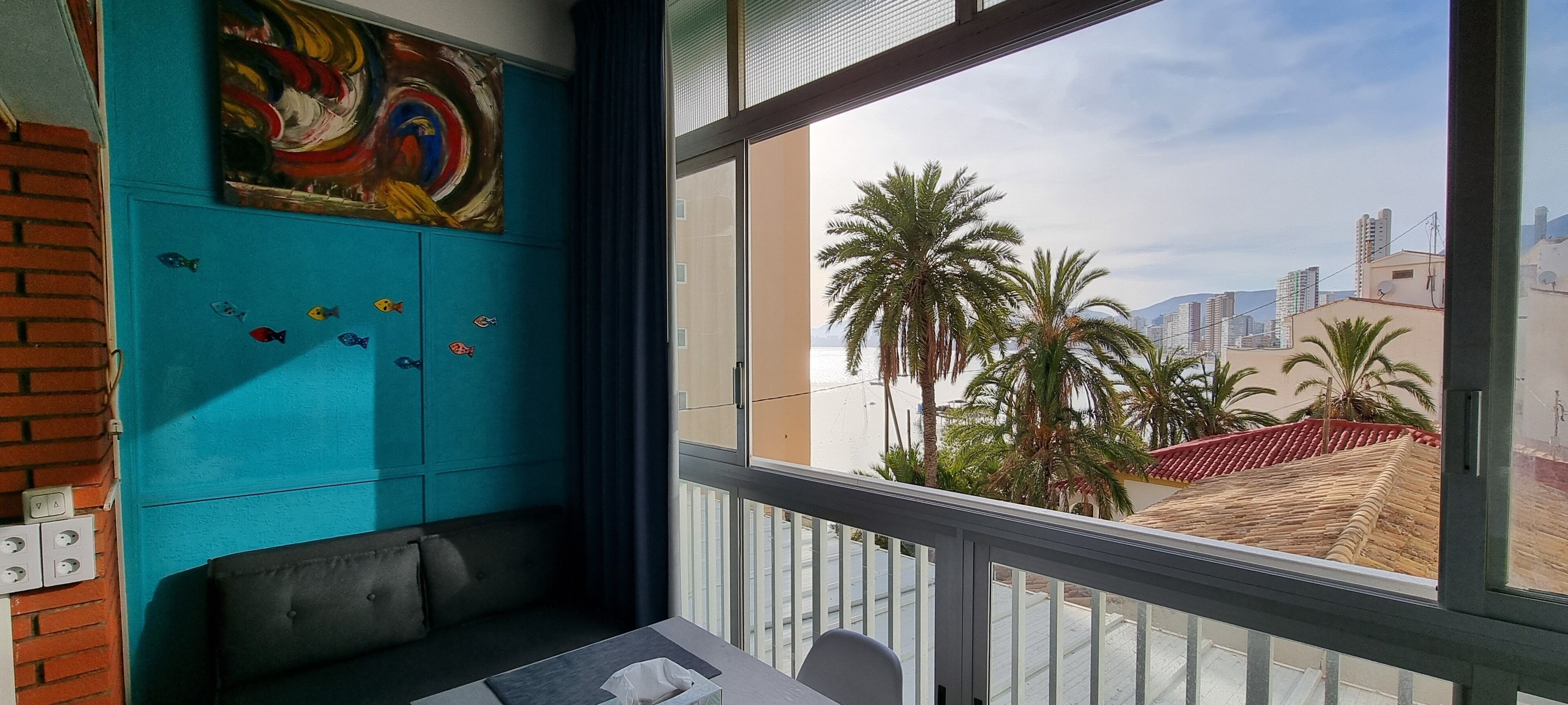 A room with a teal wall, a colorful abstract painting, a window with a view of palm trees and buildings, and a small table with a chair.