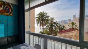 A room with a teal wall, a colorful abstract painting, a window with a view of palm trees and buildings, and a small table with a chair.