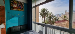 A room with a teal wall, a colorful abstract painting, a window with a view of palm trees and buildings, and a small table with a chair.