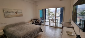 A bedroom with a bed, a desk, a chair, a balcony with a table and chairs, and a painting on the wall.