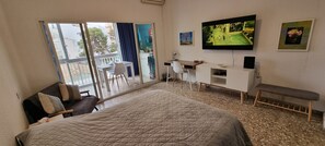 A hotel room with a bed, a desk, a TV, and a balcony with a view of buildings and trees.