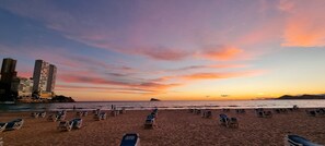 A beach with rows of sun loungers, a sunset, and buildings in the background.