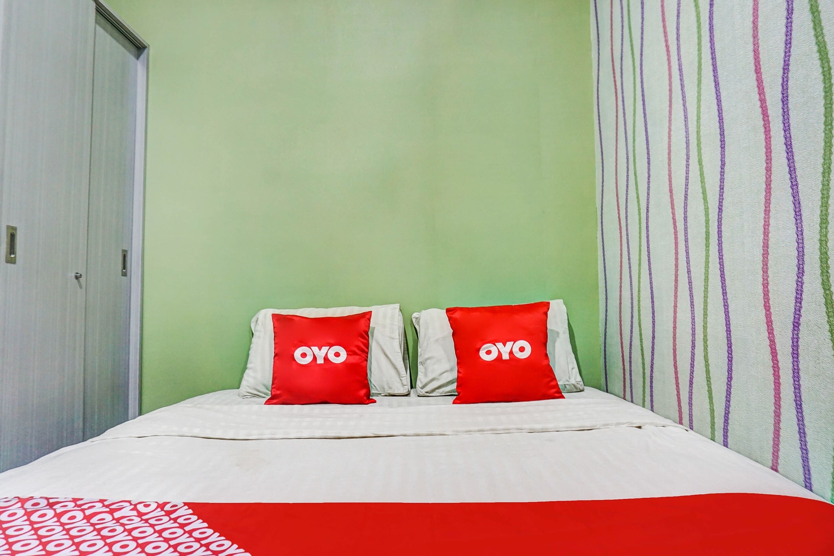 Hotellihuone, jossa on sänky, kaksi punaista tyynyä, joissa on «OYO»-merkintä, vihreä seinä ja raidallinen verho.
