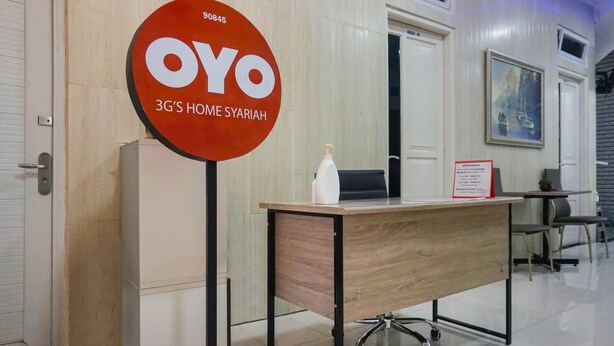 Una recepción con un letrero que dice "OYO 3G'S HOME SYARIAH" y una pequeña mesa con una botella y un cartel.