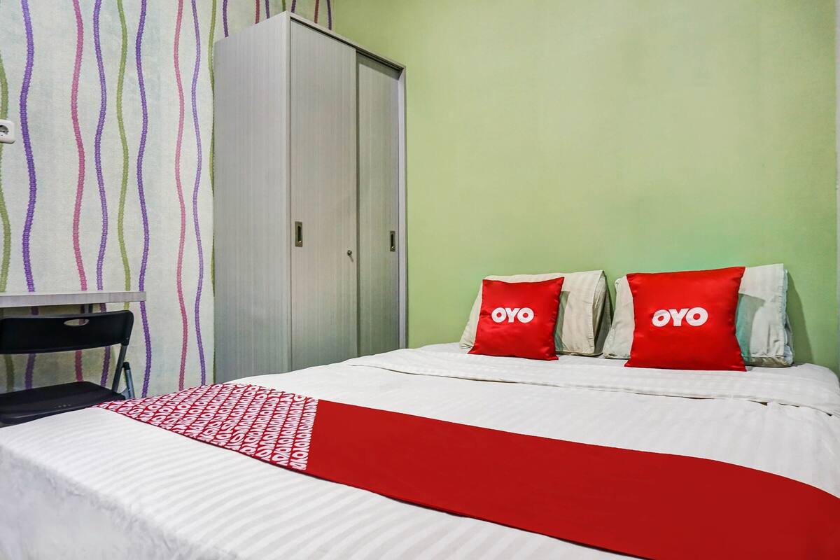 Una habitación de hotel con una cama, dos almohadas rojas con el texto "OYO", una colcha blanca y una pared a rayas.