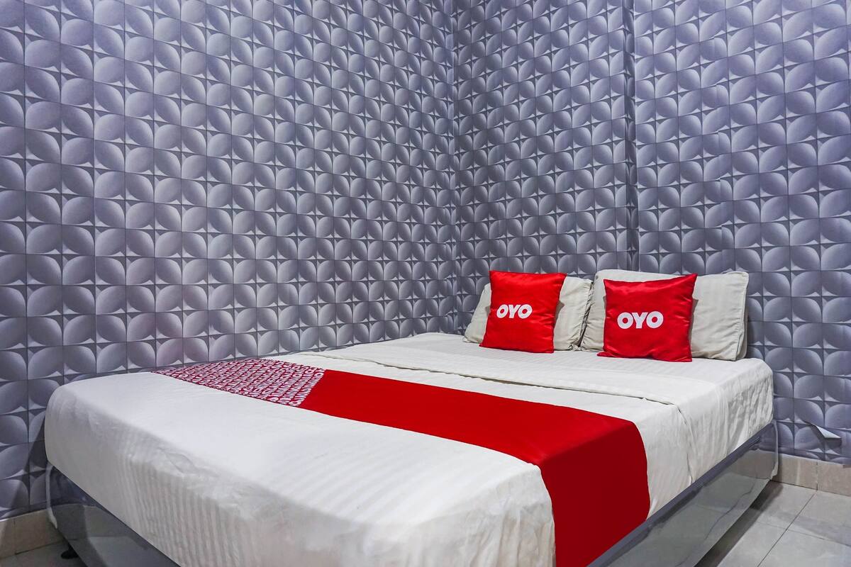 Una habitación de hotel con papel tapiz estampado, una cama con ropa blanca y detalles rojos, y dos almohadas con el nombre de marca 'OYO'.