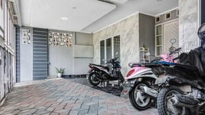 Un área de estacionamiento techada con dos motocicletas, piso de baldosas y una puerta blanca que da a una habitación con pared de azulejos y una planta en maceta.