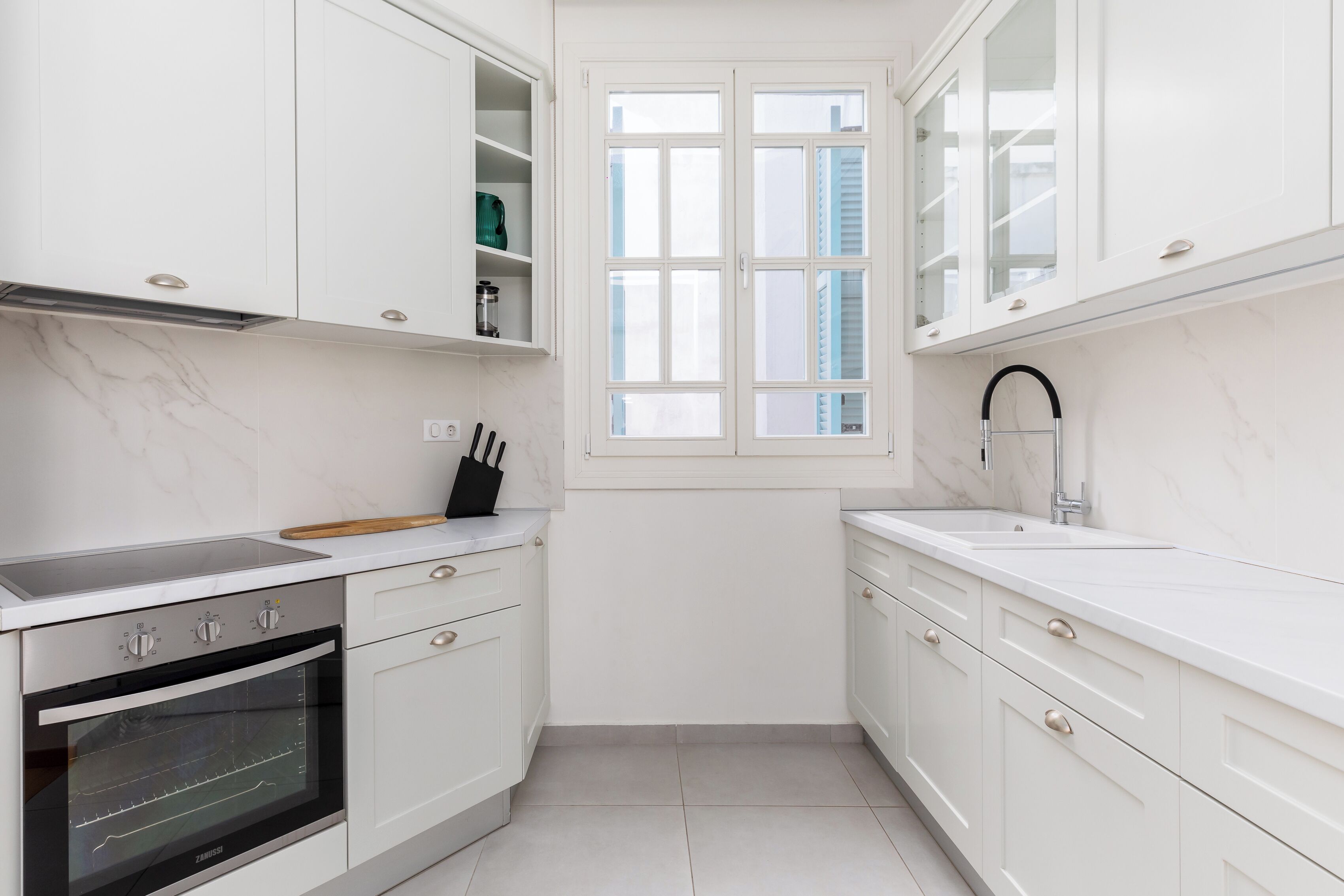 A modern kitchen with white cabinetry, a built-in oven, and a window with blinds.