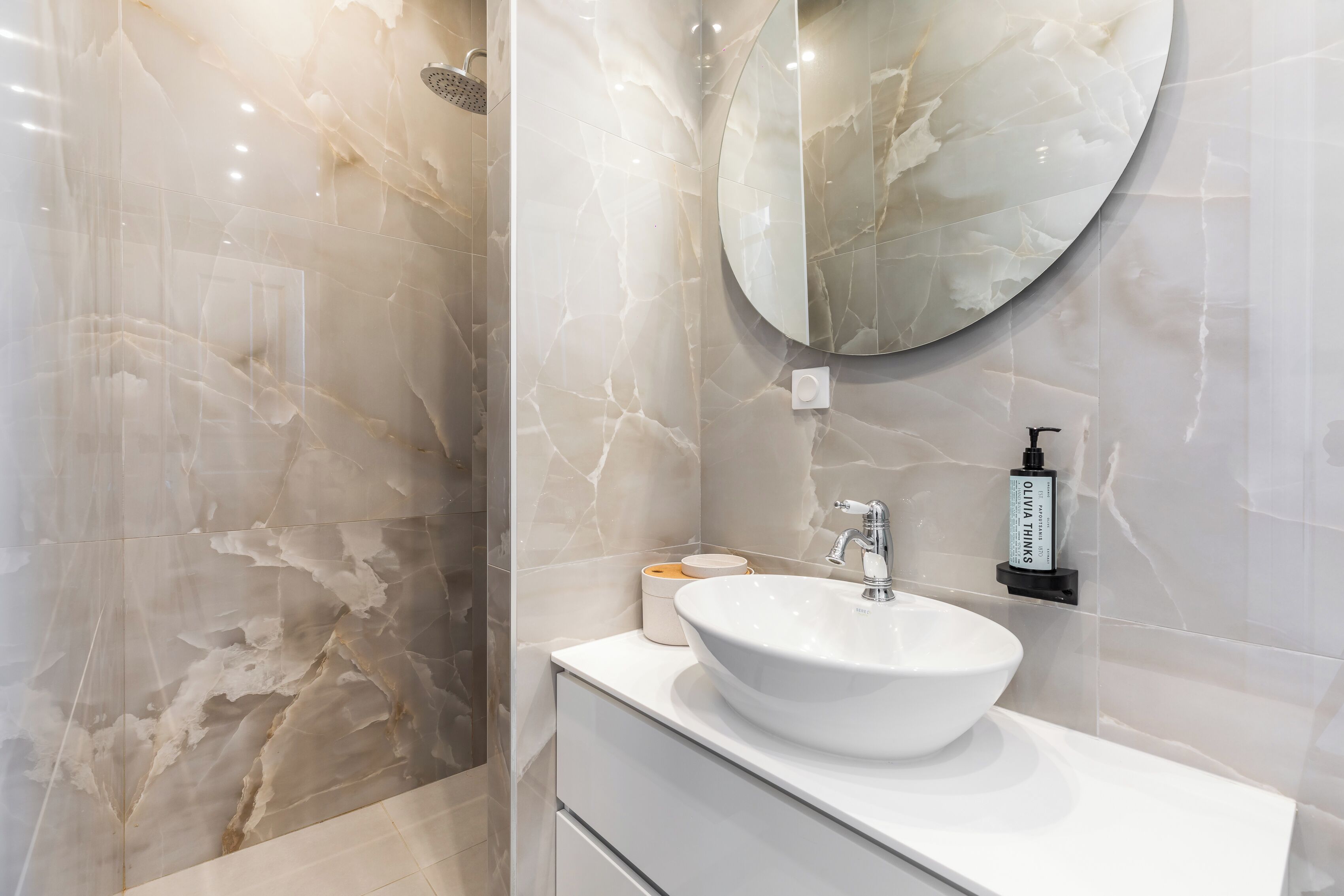 A bathroom with a white marble sink, a round mirror, and a shower area.