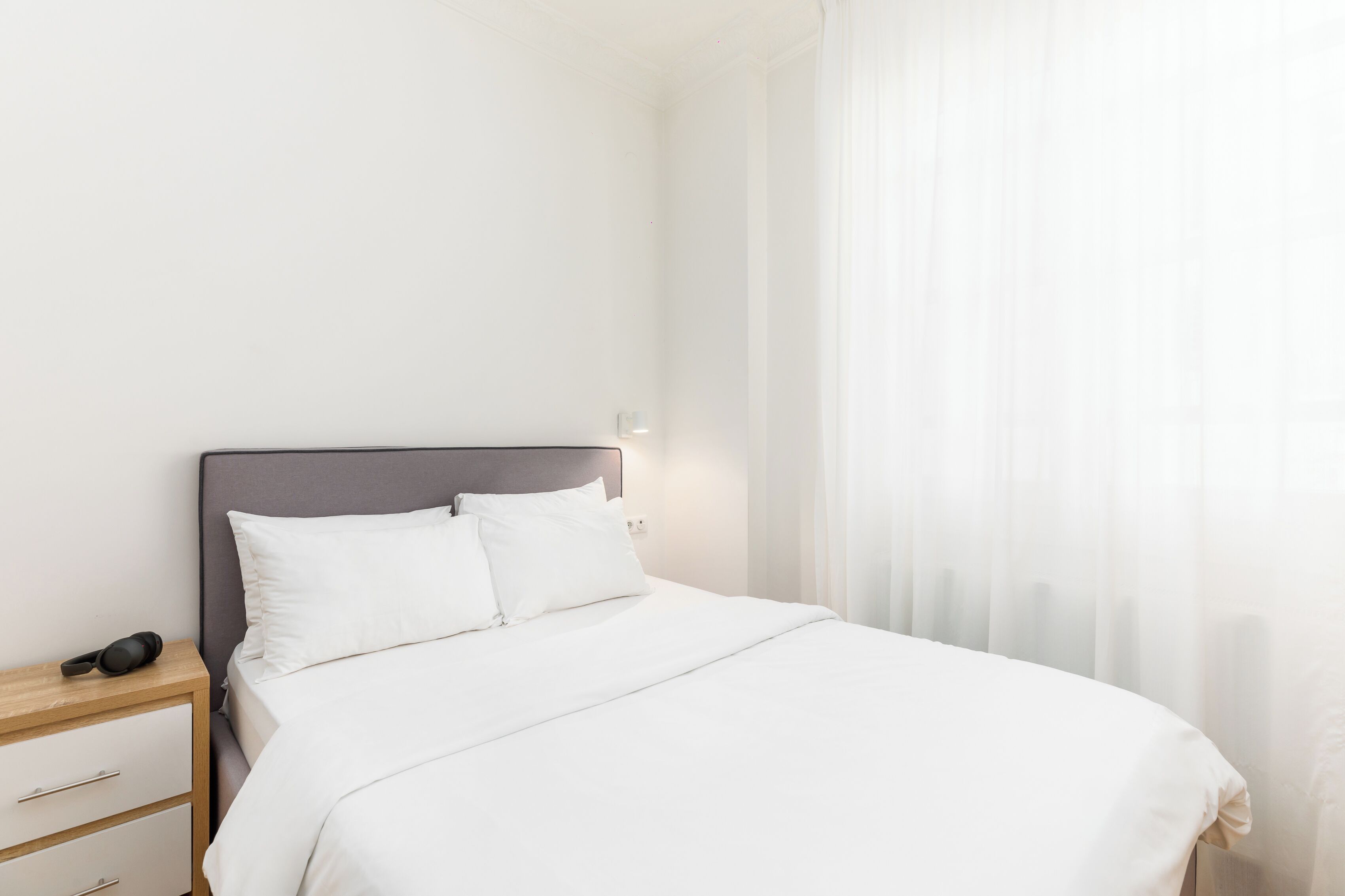 A neatly made bed with white linens, a wooden nightstand with a lamp, and a window with sheer curtains.