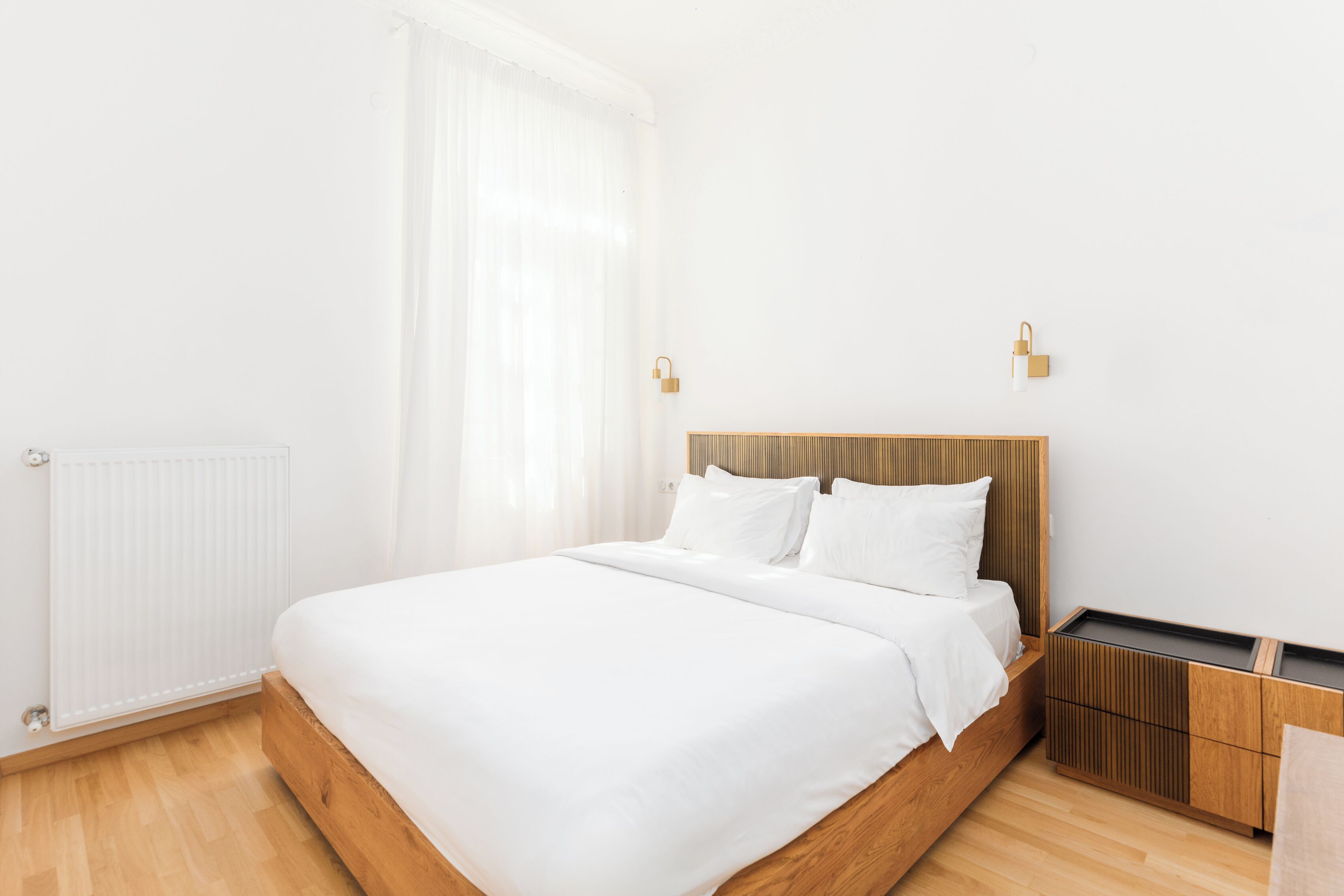 A modern bedroom with a wooden bed, white bedding, a radiator, a wooden nightstand, and a small wooden cabinet.