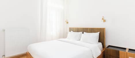 A modern bedroom with a wooden bed, white bedding, a radiator, a wooden nightstand, and a small wooden cabinet.