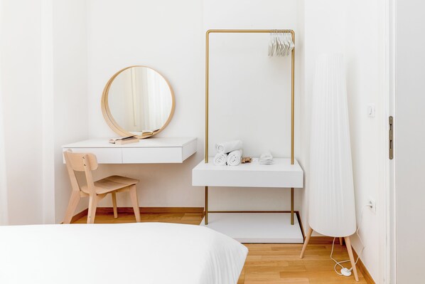 Un dormitorio minimalista con una silla de madera, un espejo redondo, un escritorio blanco y una cama blanca.