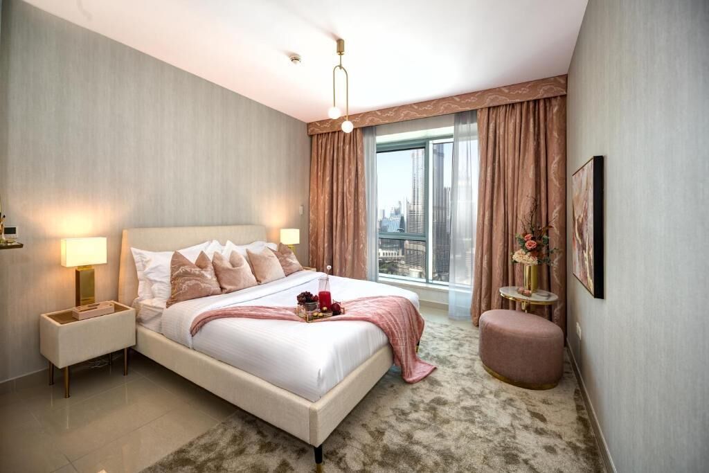 Ett modernt hotellrum med en stor säng, två sänglampor, ett litet runt bord, en rosa pall och ett stort fönster med gardiner.