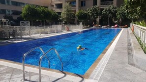 A rectangular swimming pool with a metal ladder in the foreground, surrounded by a tiled deck and a white fence. - Durrani Homes - Designer 2BR Apt with stunning Burj Khalifa and Fountain View (Dubai)