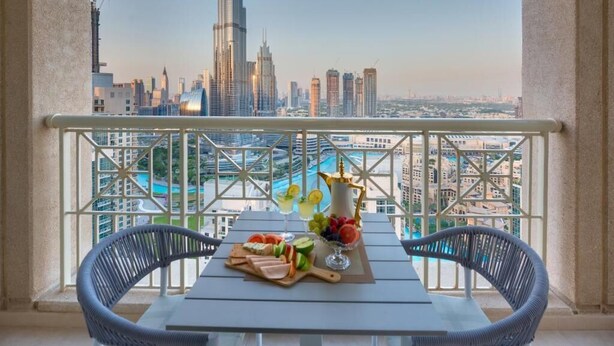 A balcony with a table set for a meal, featuring a fruit platter and a decorative vase. The view includes a city skyline with a prominent skyscraper. - Durrani Homes - Designer 2BR Apt with stunning Burj Khalifa and Fountain View (Dubai)