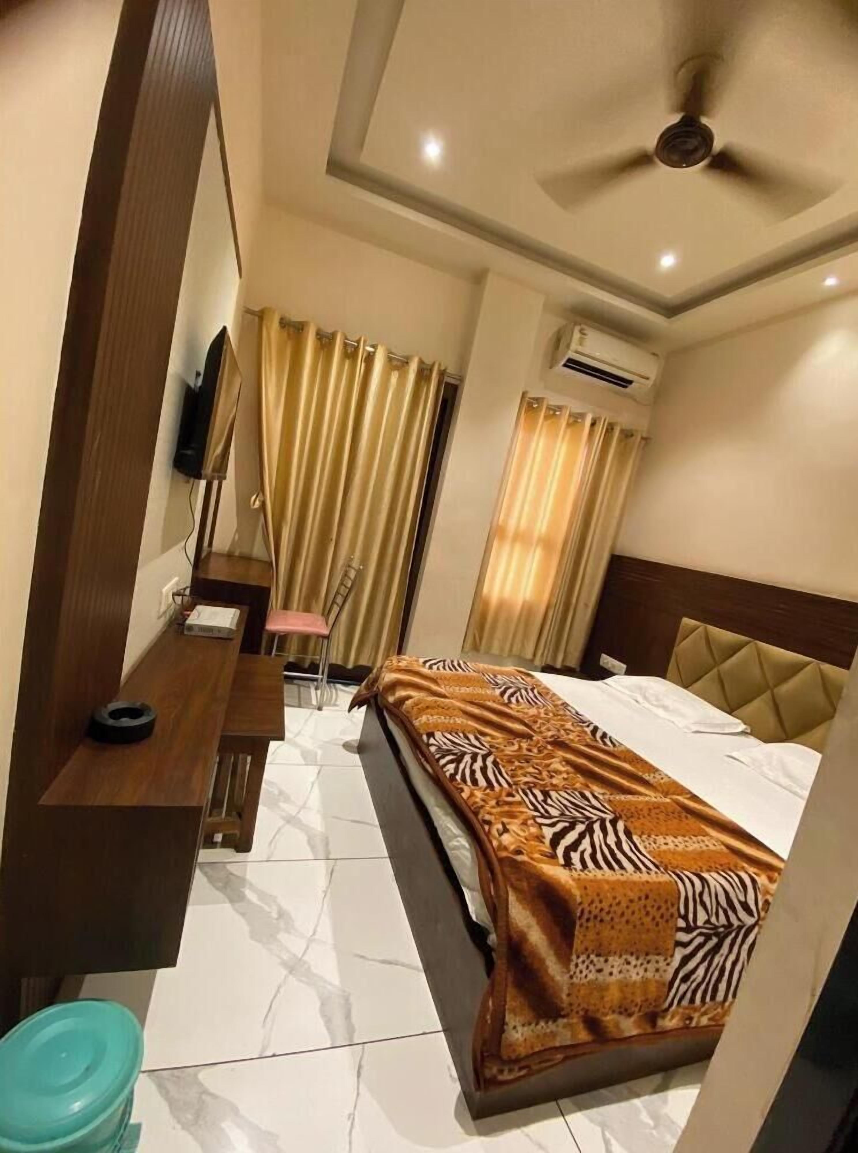 Habitación de hotel con cama, escritorio, silla, televisor, cortinas, unidad de aire acondicionado y ventilador de techo.