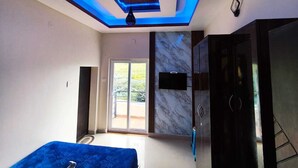 A modern bedroom with a ceiling light fixture, a bed with a blue cover, a TV mounted on the wall, and a sliding door leading to an outdoor area. - Rose garden enclave for families  (Ooty)
