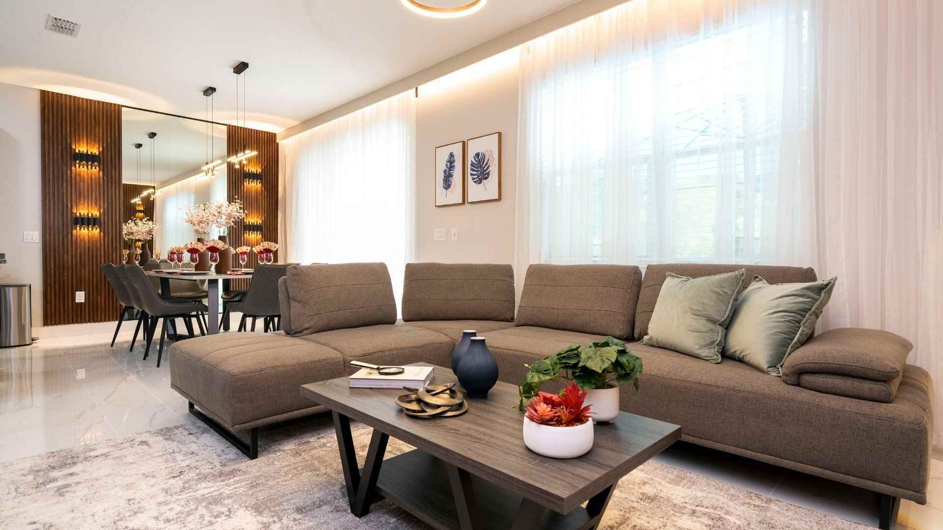 Un salon moderne comprenant un canapé d’angle, une table basse agrémentée d’objets décoratifs, un coin repas avec un lustre et de grandes fenêtres ornées de rideaux légers.