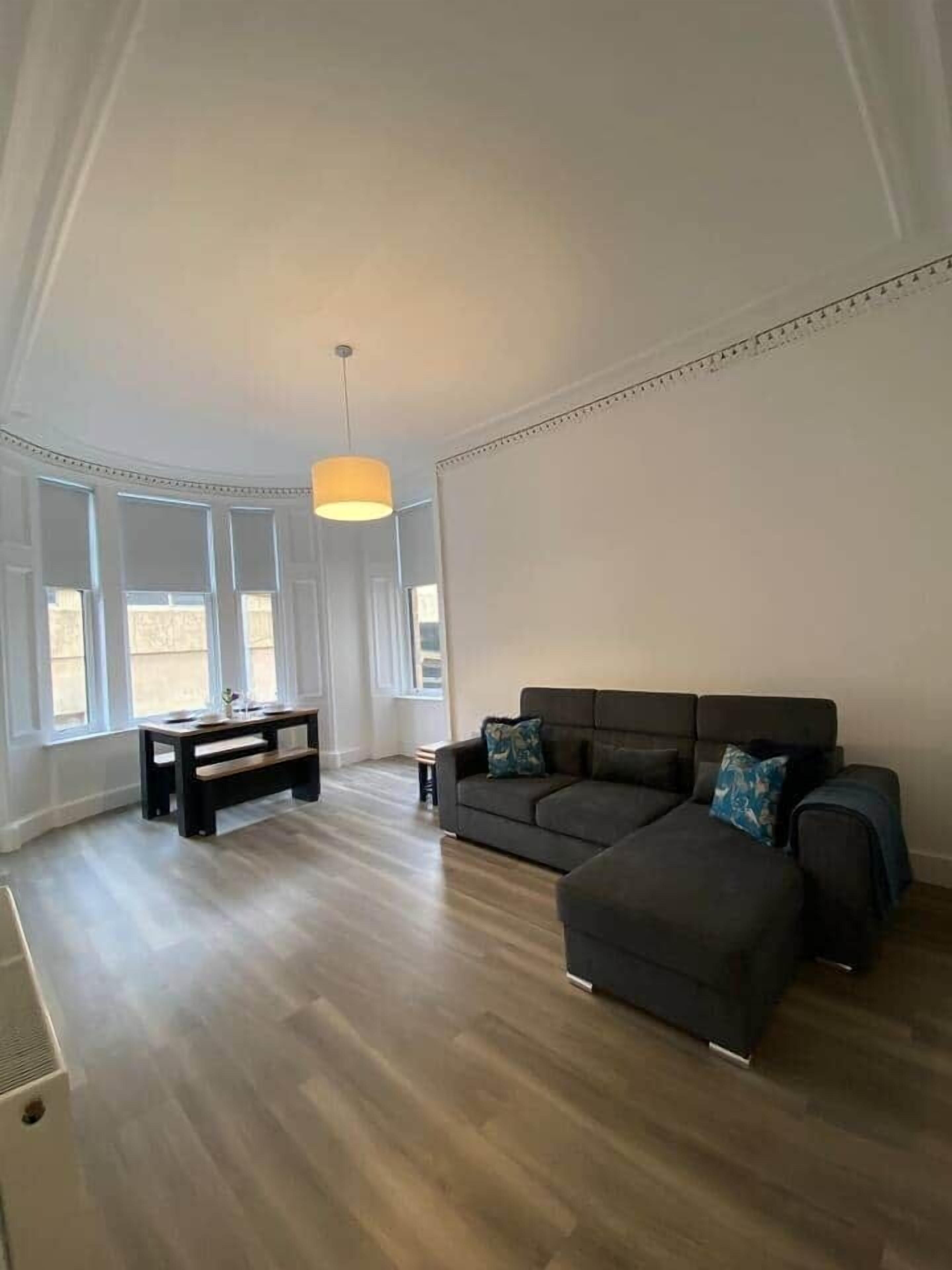 A spacious living room with a dark grey sectional sofa, a small black coffee table, and a bench. Large windows allow natural light to fill the room.