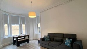 Een ruime woonkamer met een donkergrijze hoekbank, een klein zwart salontafeltje en een bankje. Grote ramen laten natuurlijk licht de kamer binnen.