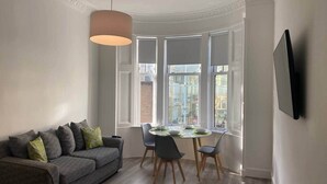 Una sala de estar moderna con un sofá gris, una mesa de comedor con sillas y un amplio ventanal con múltiples cristales.