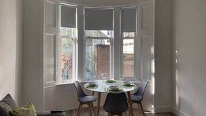 Un comedor con una mesa redonda para cuatro personas, un ventanal con marcos blancos y un sofá gris con un cojín verde.