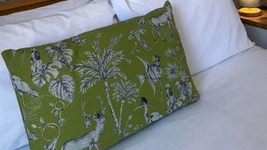 Una cama con sábanas blancas y almohada verde con estampado floral, frente a una pared con diseño de estampado paisley.