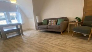 Un salon avec un plancher en bois, un canapé gris agrémenté de coussins verts, une petite table et une chaise.