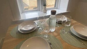 Une table à manger, dressée avec des assiettes, des verres et des couverts, est placée près d’une fenêtre d’où l’on peut admirer la vue sur les bâtiments.