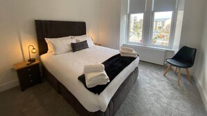 Una cama bien hecha con sábanas blancas y un plaid negro, una lámpara de noche y una silla junto a la ventana.