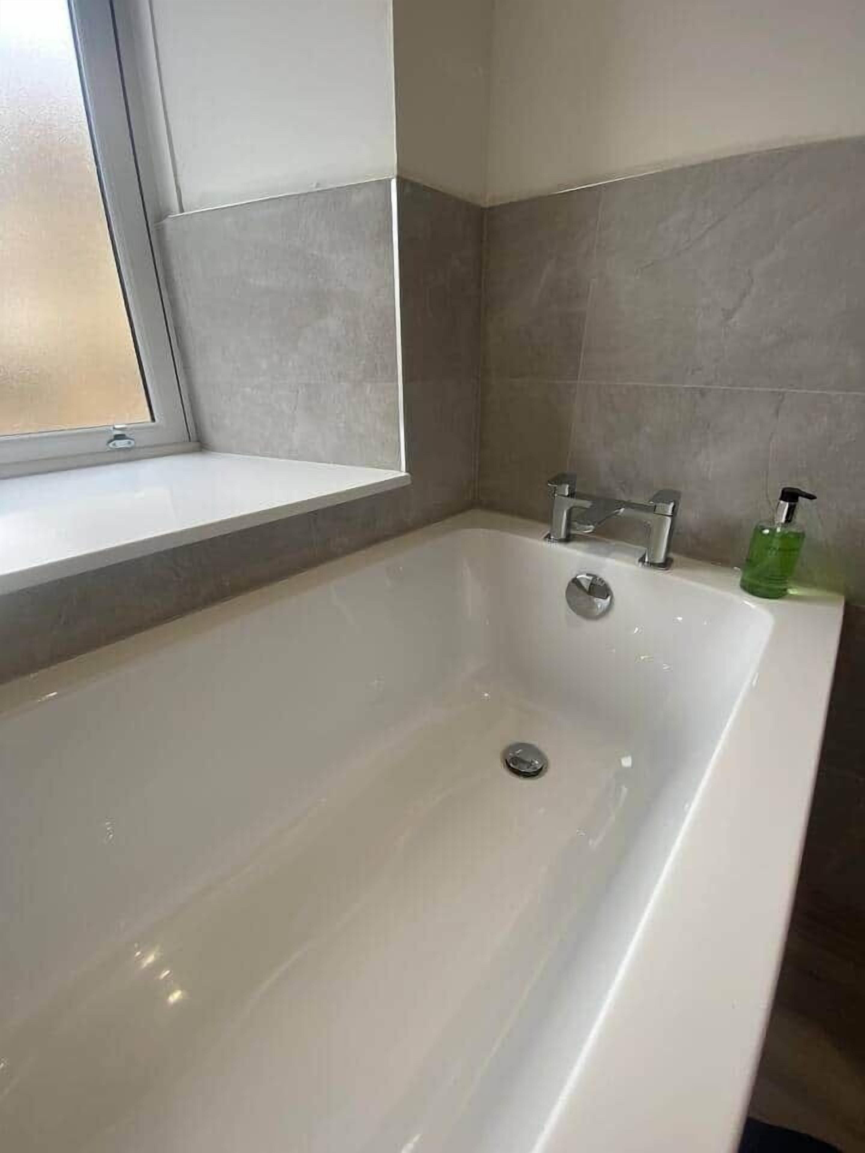 A modern bathroom with a freestanding bathtub, a window, and a green soap dispenser.