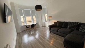 Amplia sala de estar con un sofá gris oscuro, una mesa de comedor con sillas y un ventanal que proporciona luz natural.