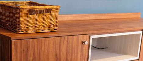 Ein Holzschrank mit einem Korbwäsche darüber, einem darüber angebrachten Fernseher und einem grauen Sessel mit Decke.