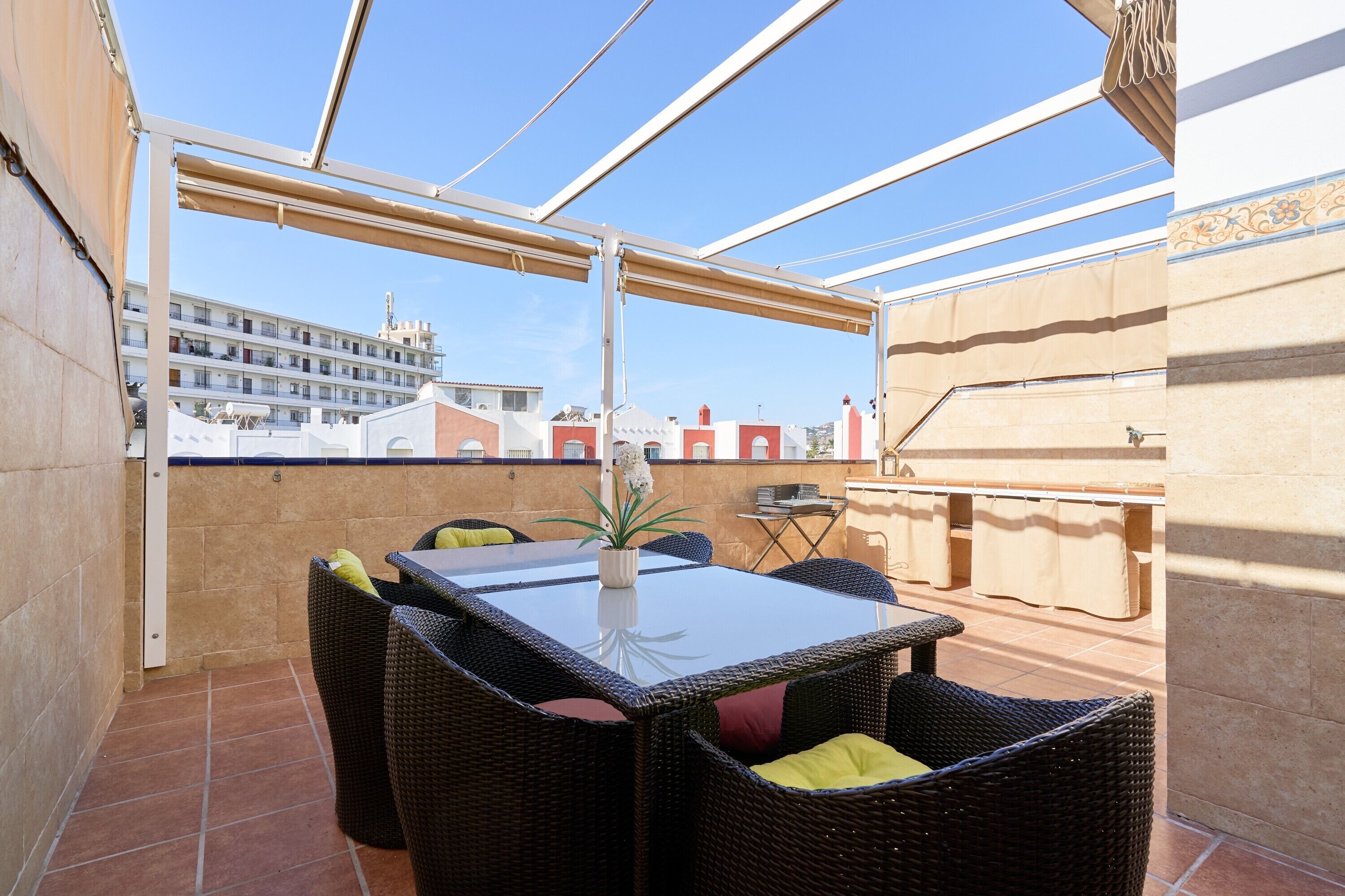 A terrace with a table and chairs, a potted plant, and a view of buildings in the background.