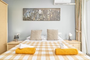 A hotel room with a checkered bedspread, two pillows, a cityscape artwork on the wall, a bedside lamp, and a window with curtains.