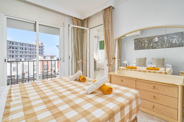 A hotel room with a checkered bedspread, a large mirror, a cityscape view, and a balcony with curtains.