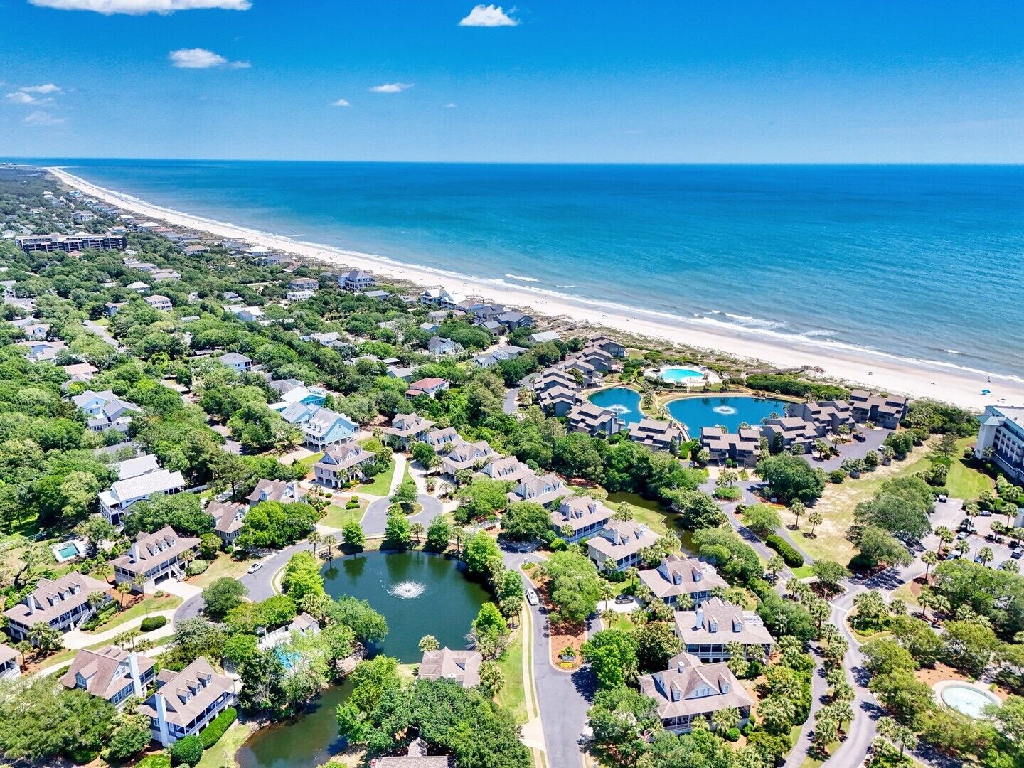 An aerial view of a coastal residential area with houses, a beach, and a resort with swimming pools.