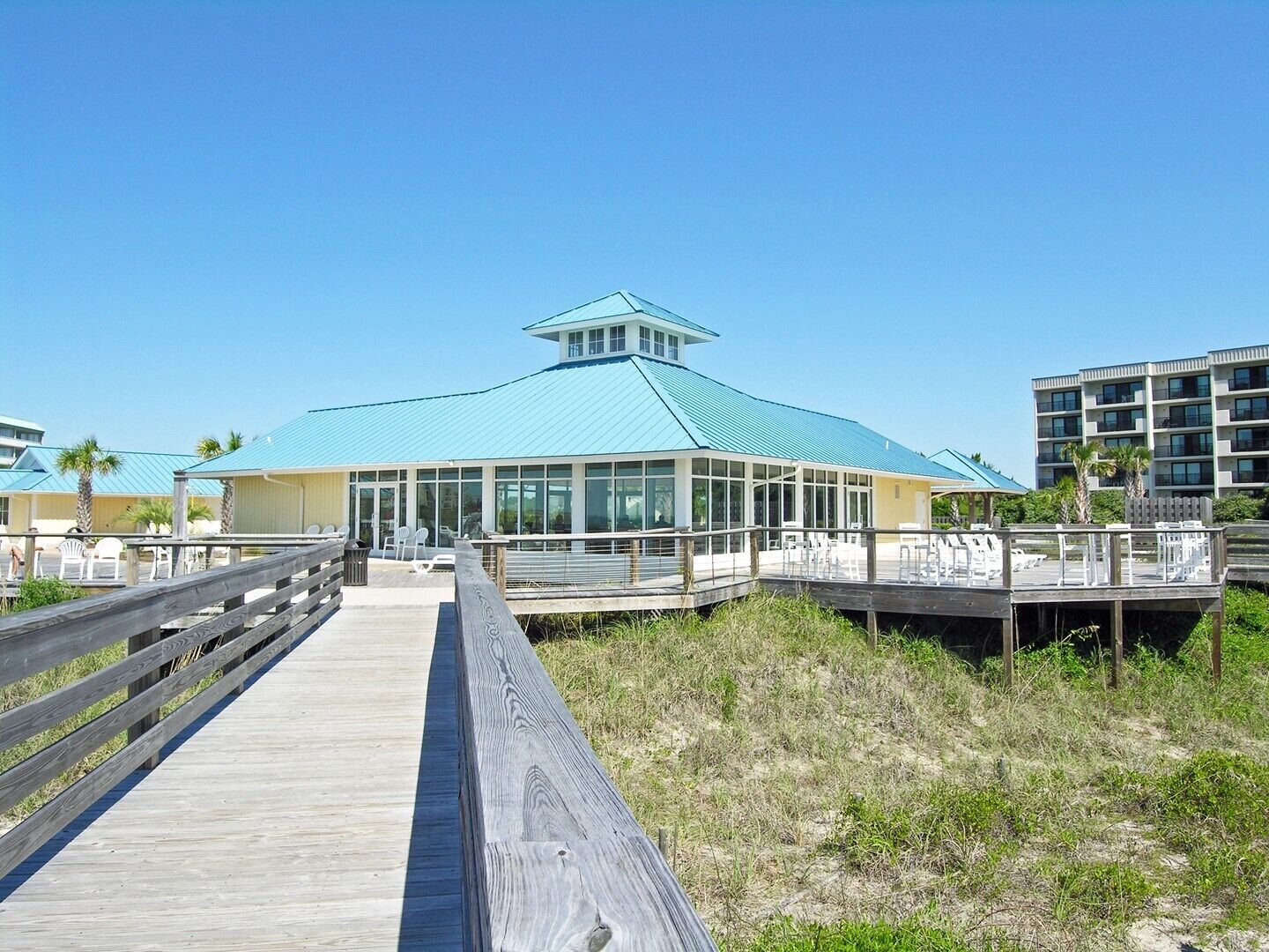 A coastal building with a blue roof, a glass facade, and a wooden boardwalk leading to it.