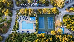 An aerial view of a property with a swimming pool, tennis courts, and a parking area surrounded by trees.