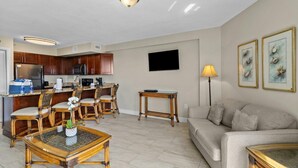 A modern living room with a sofa, a dining area with chairs, a kitchen with cabinets, a flat-screen TV, and artwork on the wall.