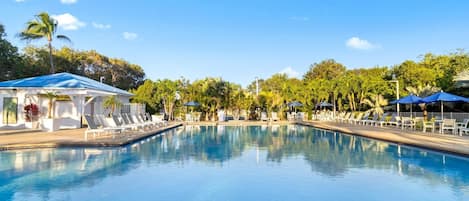 A clear swimming pool with lounge chairs and umbrellas, surrounded by palm trees and a clear sky.