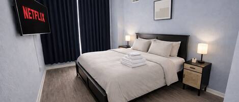 A modern hotel room with a bed, two bedside tables with lamps, a TV mounted on the wall, and a window with curtains.