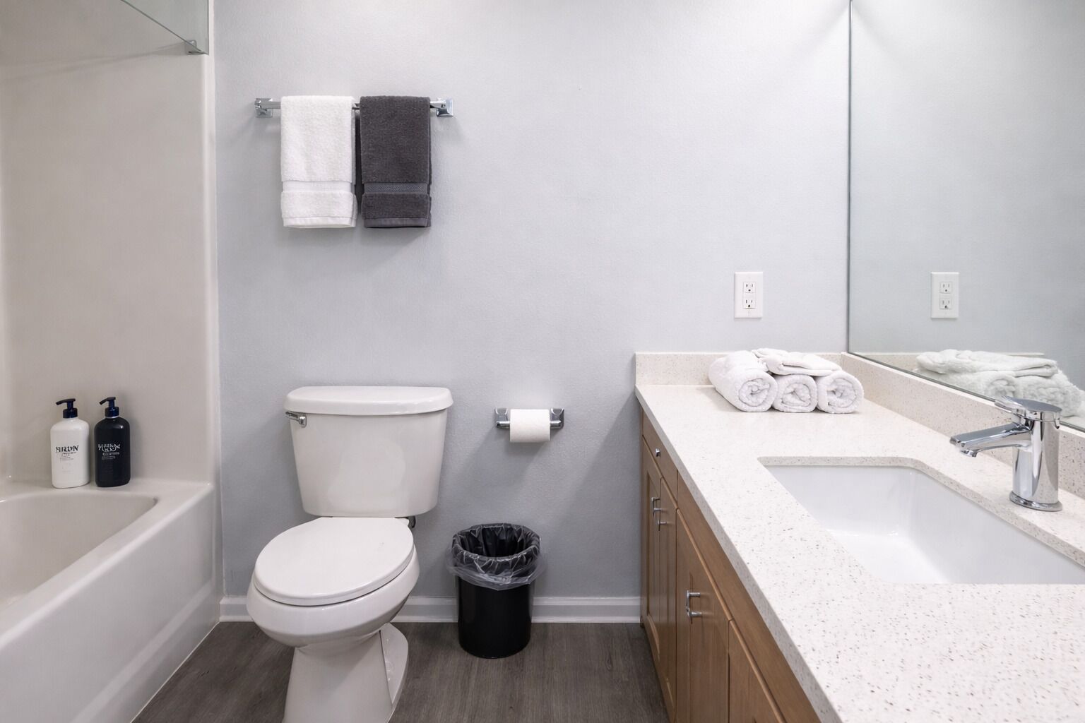 A modern bathroom with a toilet, sink, and mirror. There are towels on the wall and a trash can.