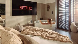 A modern bedroom with a large bed, a TV displaying 'NETFLIX', a bathtub, and a large window with blinds. - Granaria Jacuzzi Premium Suites by Downtown Apartments (Gdańsk)