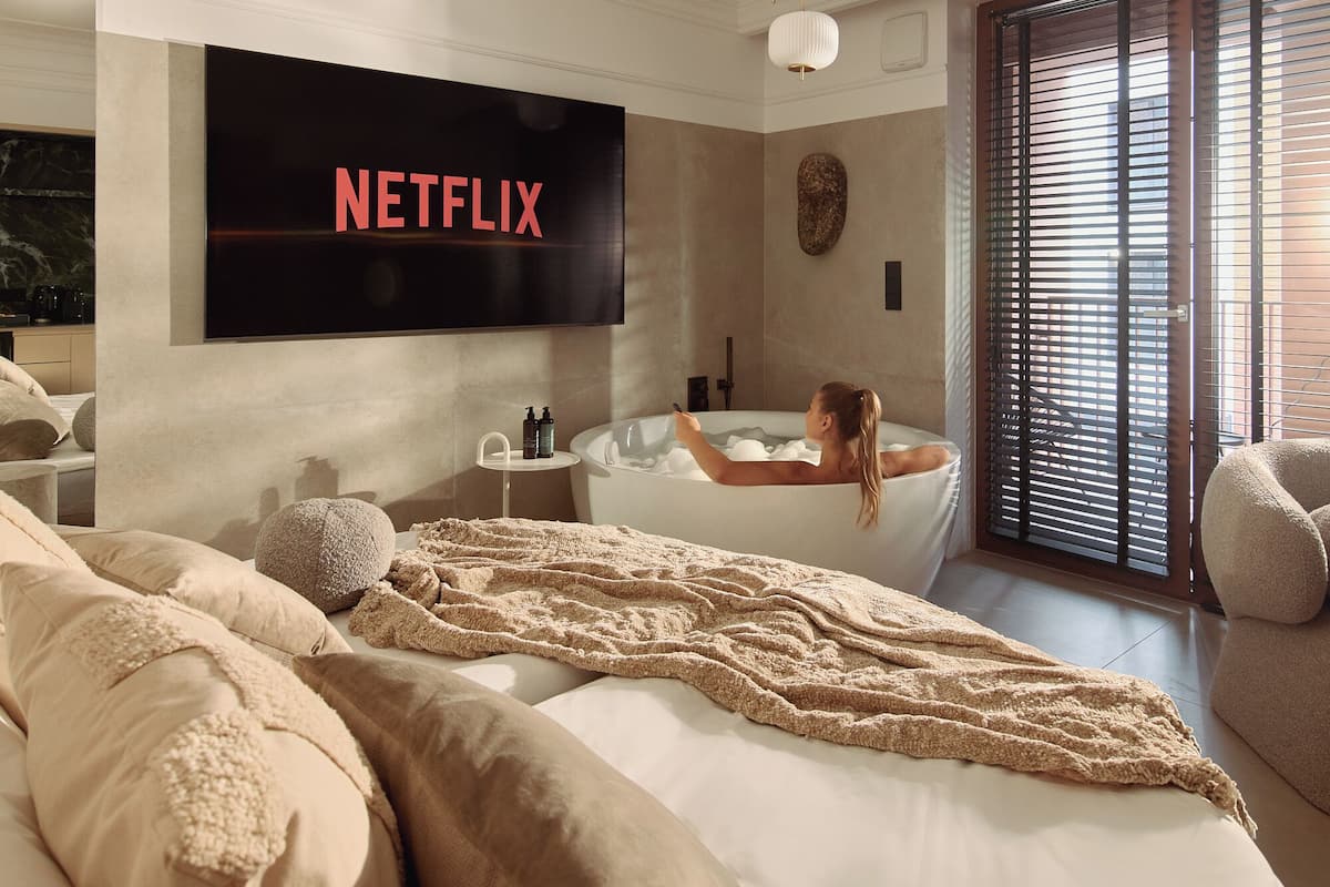 A modern bedroom with a large bed, a TV displaying 'NETFLIX', a bathtub, and a large window with blinds.