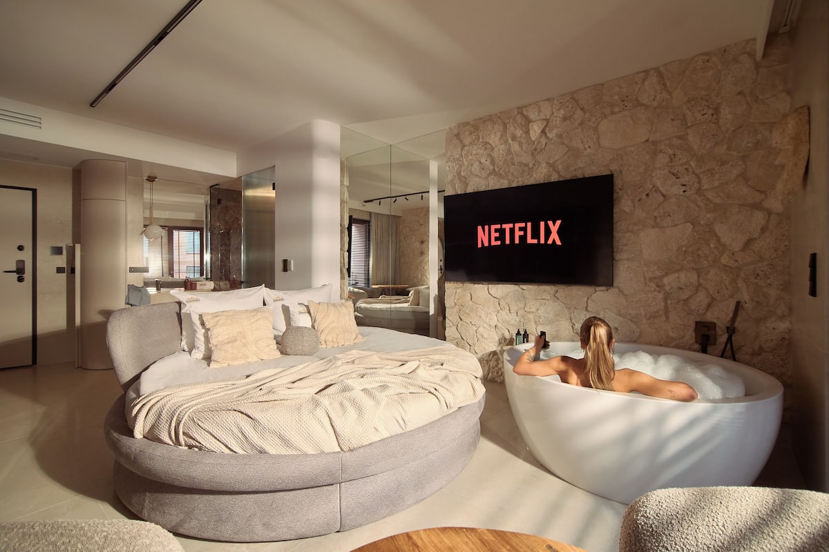 A modern bedroom with a large bed, a bathtub, and a TV displaying 'NETFLIX'.